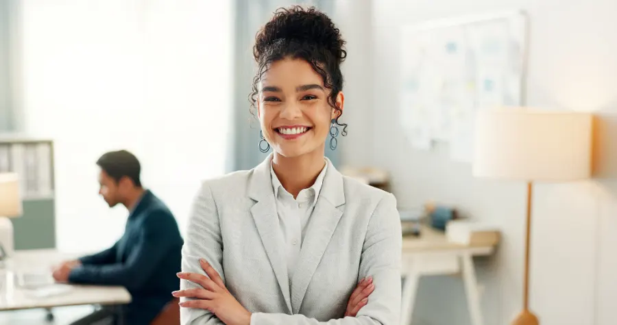 A young woman in a suit stands in the office, smiling at the camera.