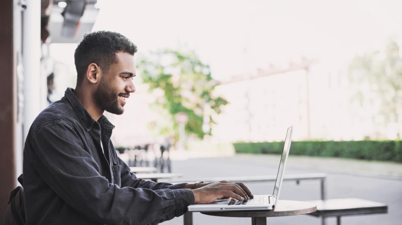 A young man sits outside, working on his laptop.